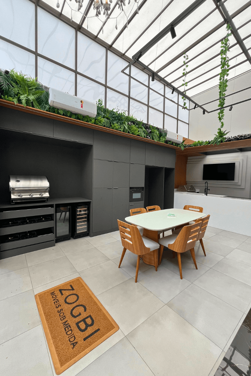 Área gourmet de luxo com teto de vidro, armários planejados cinza chumbo, churrasqueira inox e decoração com plantas no teto.