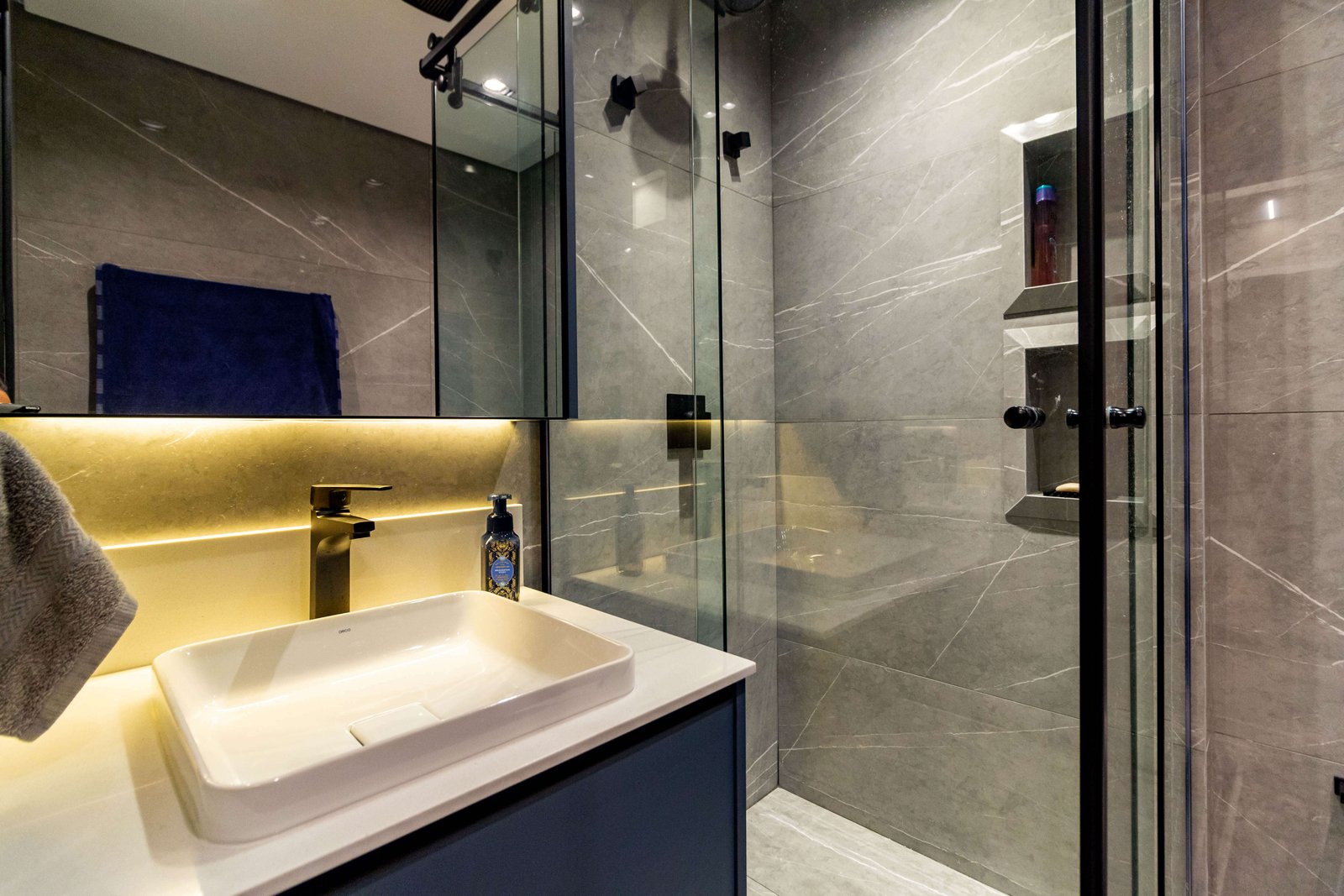 Banheiro planejado com gabinete azul marinho, metais pretos foscos, espelho com iluminação LED e revestimento cinza marmorizado.