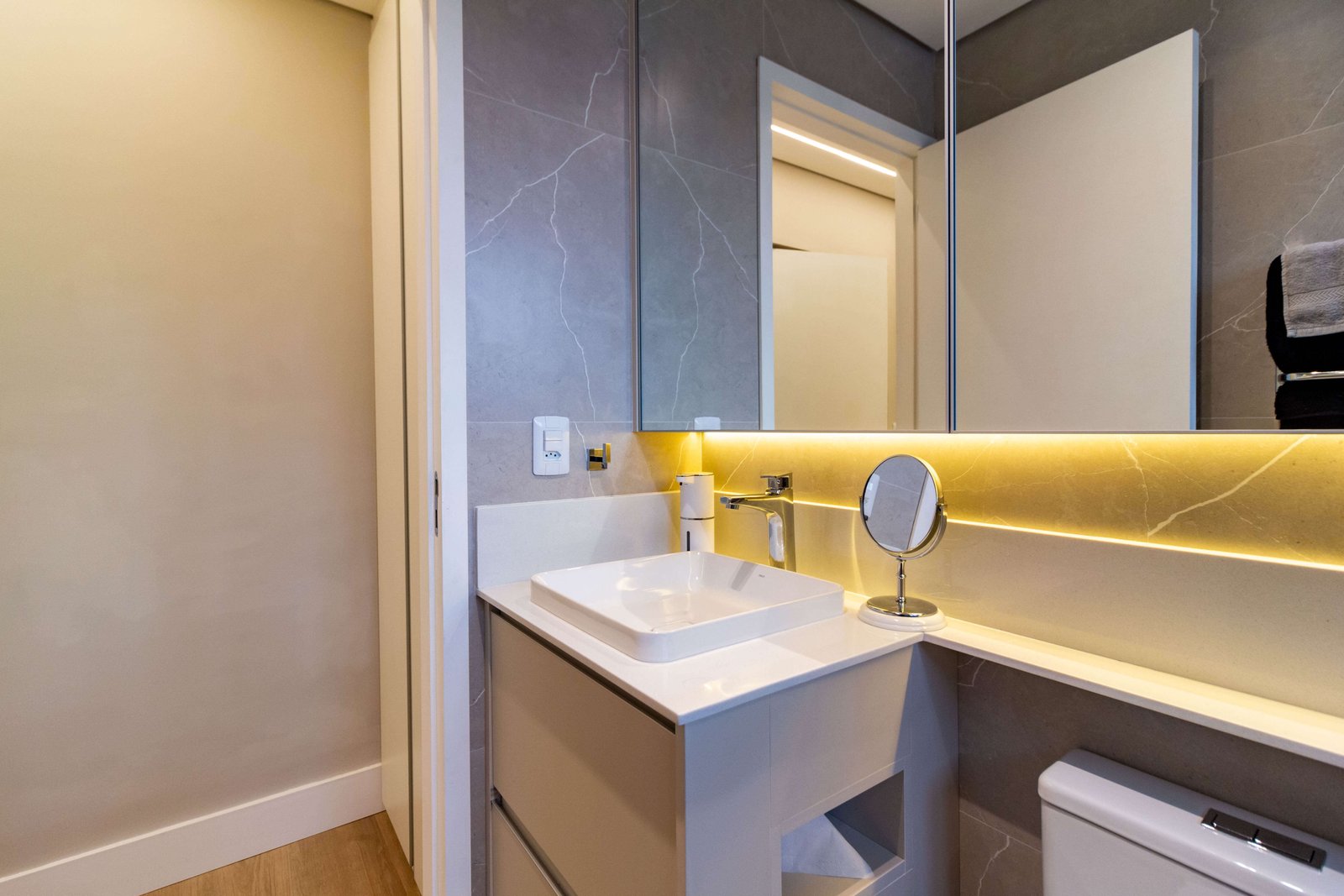 Banheiro planejado cinza com bancada branca, cuba de apoio quadrada e armário espelhado com iluminação LED indireta na base.