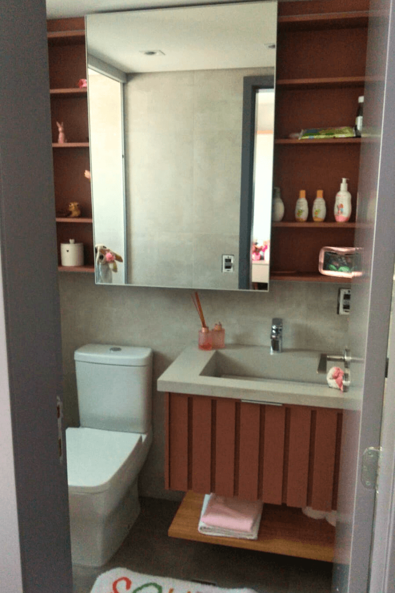 Um lavabo com gabinete inferior na cor terracota (marrom avermelhado) com detalhe ripado. Acima da bancada cinza com cuba esculpida, há um armário superior com espelho e nichos laterais abertos.