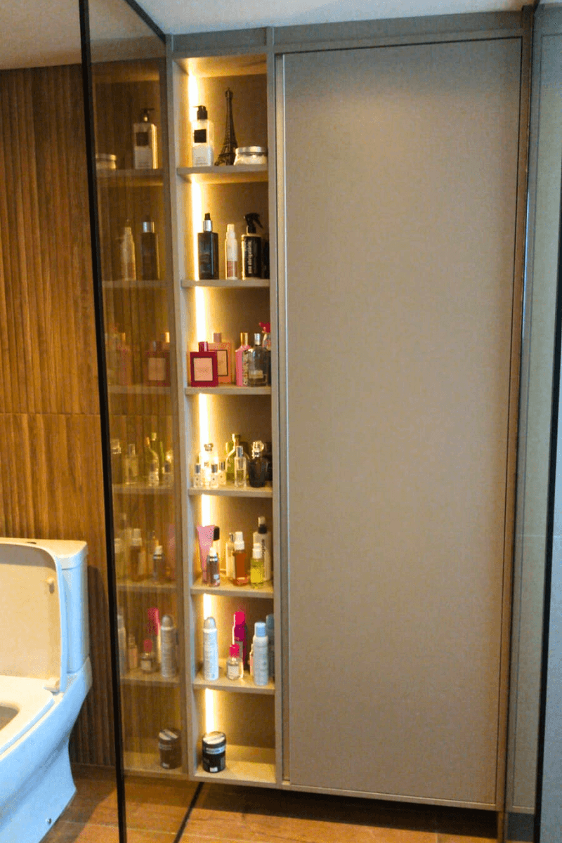 Uma torre estreita de marcenaria (roupeiro ou estante) com prateleiras iluminadas por LED vertical, exibindo uma coleção organizada de perfumes e cosméticos. O acabamento é amadeirado.