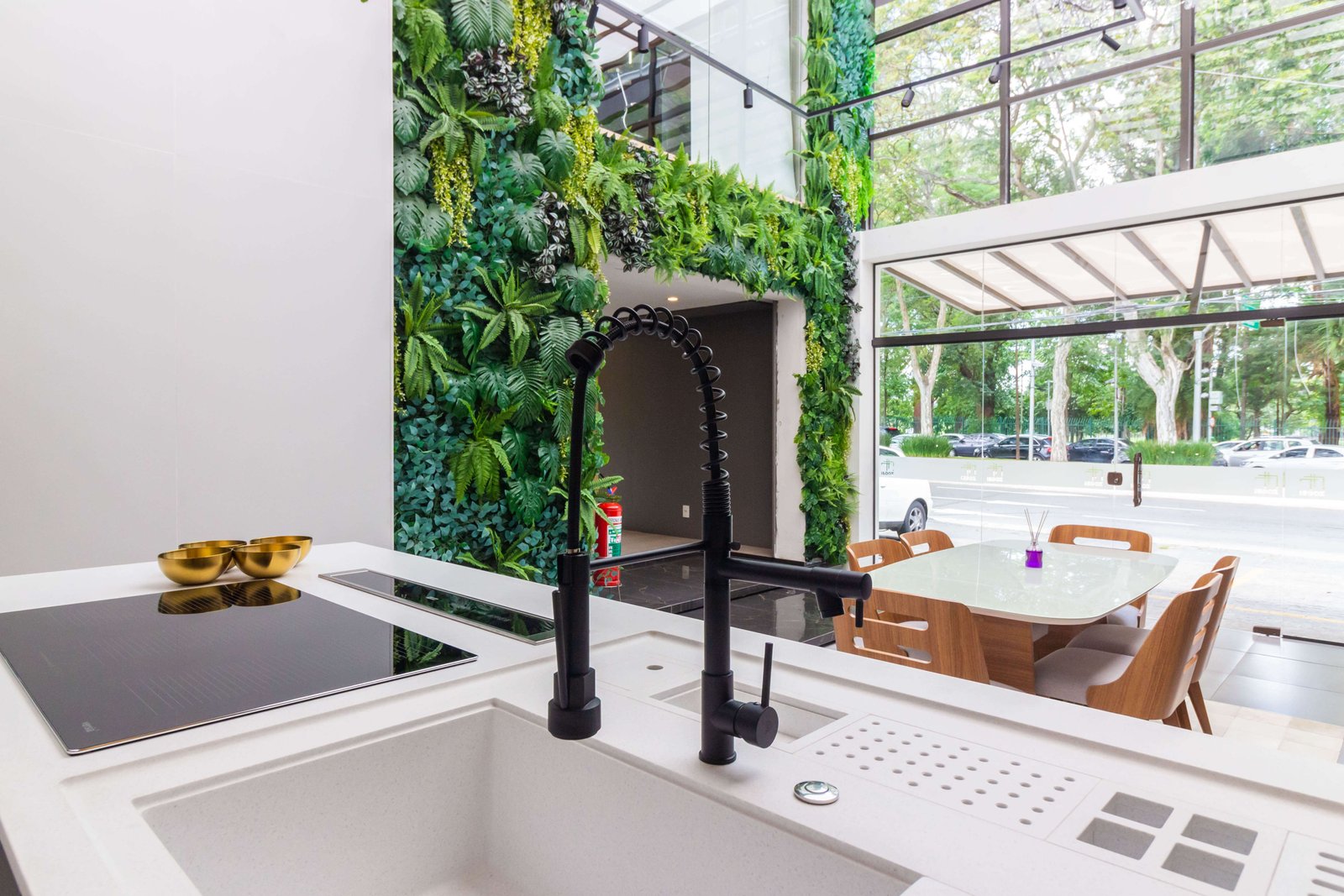 Close-up de uma pia de ilha com uma torneira gourmet preta flexível e calha úmida embutida. Ao fundo, através de grandes janelas de vidro, vê-se uma parede verde (jardim vertical).