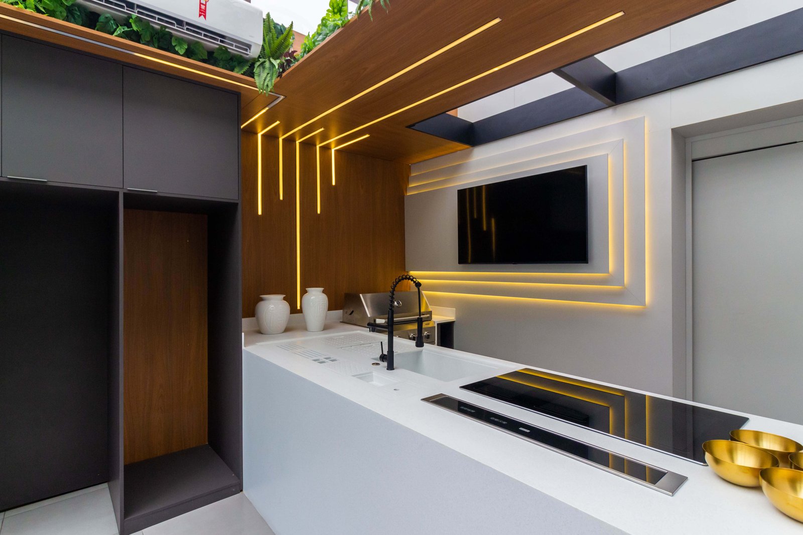 Outro ângulo da mesma área gourmet, focando no painel da TV retroiluminado e na churrasqueira a gás embutida na bancada de pedra branca.