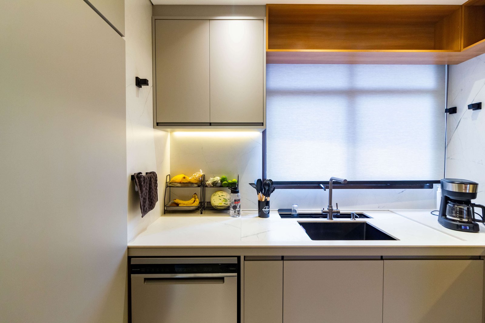 A área da pia sob uma janela com persiana branca. Acima, há um detalhe em marcenaria amadeirada no teto e, abaixo, uma lava-louças em inox embutida nos armários bege.