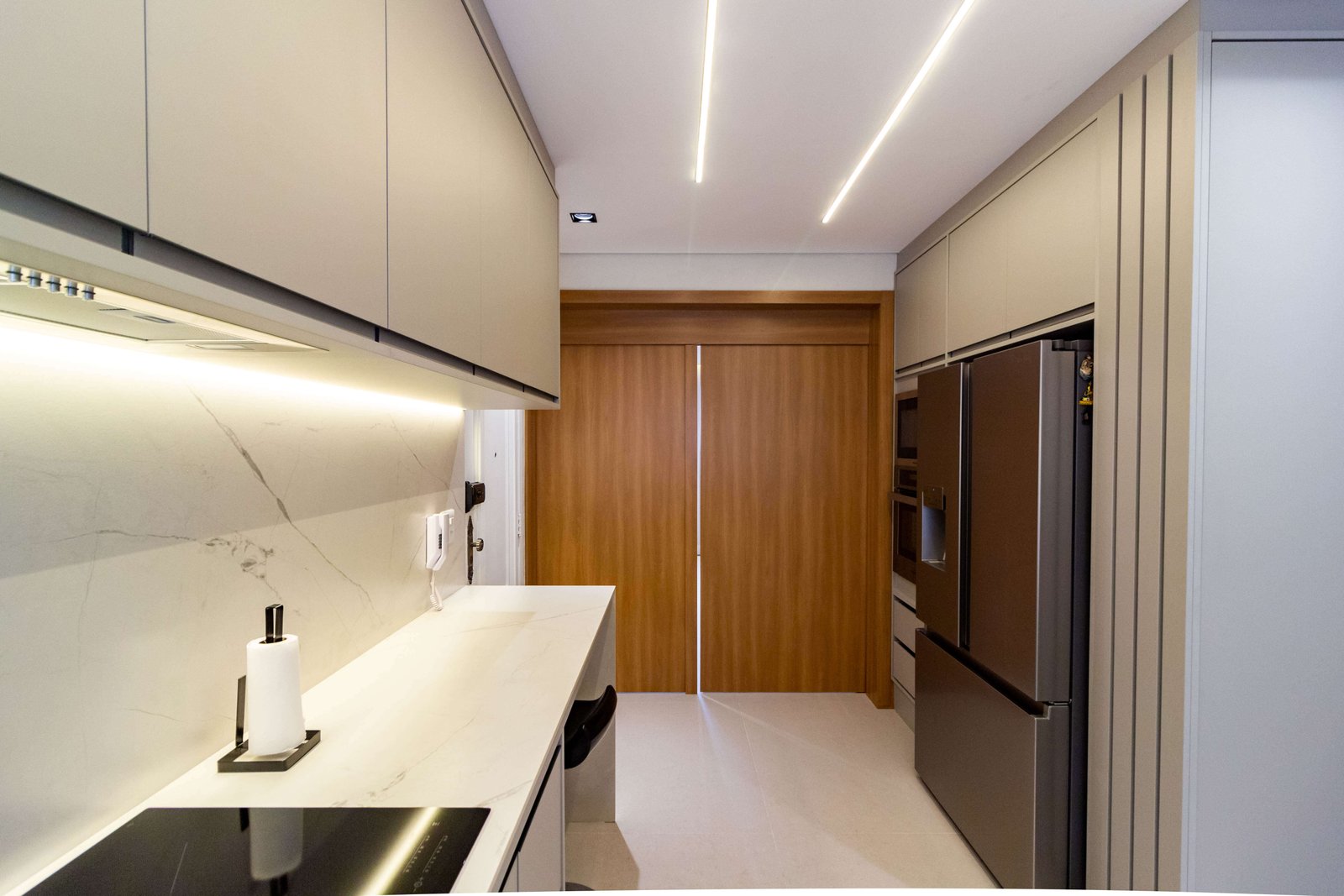 O corredor da cozinha, mostrando uma geladeira de inox grande (French Door) embutida na marcenaria à direita, e uma porta de correr de madeira ao fundo. O teto possui iluminação linear em LED.