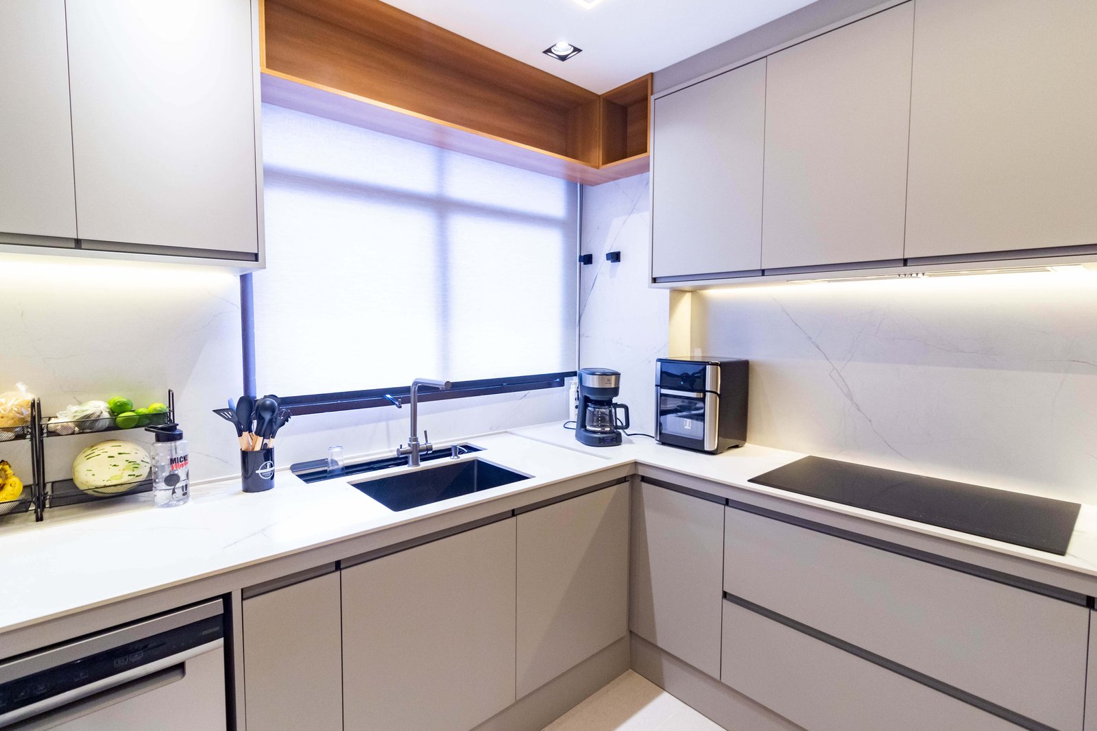 O canto da bancada em 'L', com eletroportáteis (cafeteira e air fryer) sobre a pedra branca e revestimento de parede marmorizado, iluminado por fita de LED sob o armário superior.