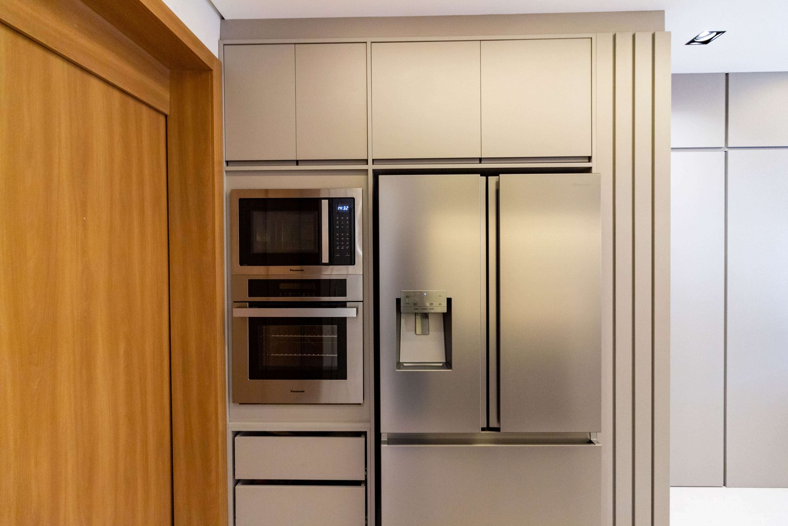 Detalhe de uma 'torre quente' planejada em tom cinza, contendo forno e micro-ondas embutidos em aço inox. Ao lado, uma geladeira side-by-side inox encaixada na marcenaria, com um detalhe ripado vertical na lateral.
