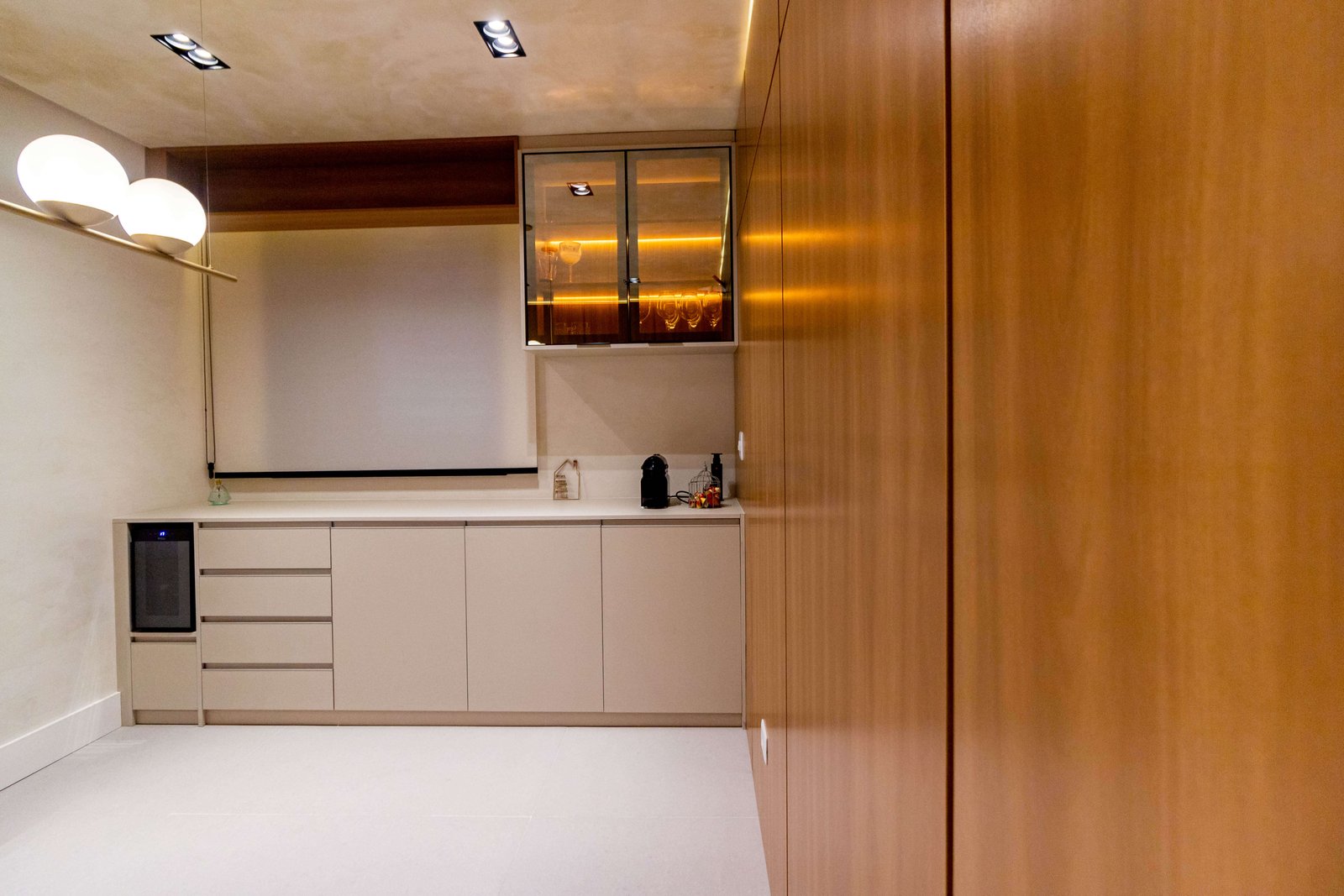 Um ambiente de copa ou buffet com armários inferiores bege e tampo claro. À direita, uma parede revestida com painel de madeira. Acima, um armário aéreo com portas de vidro reflexivo e iluminação interna amarela (quente).