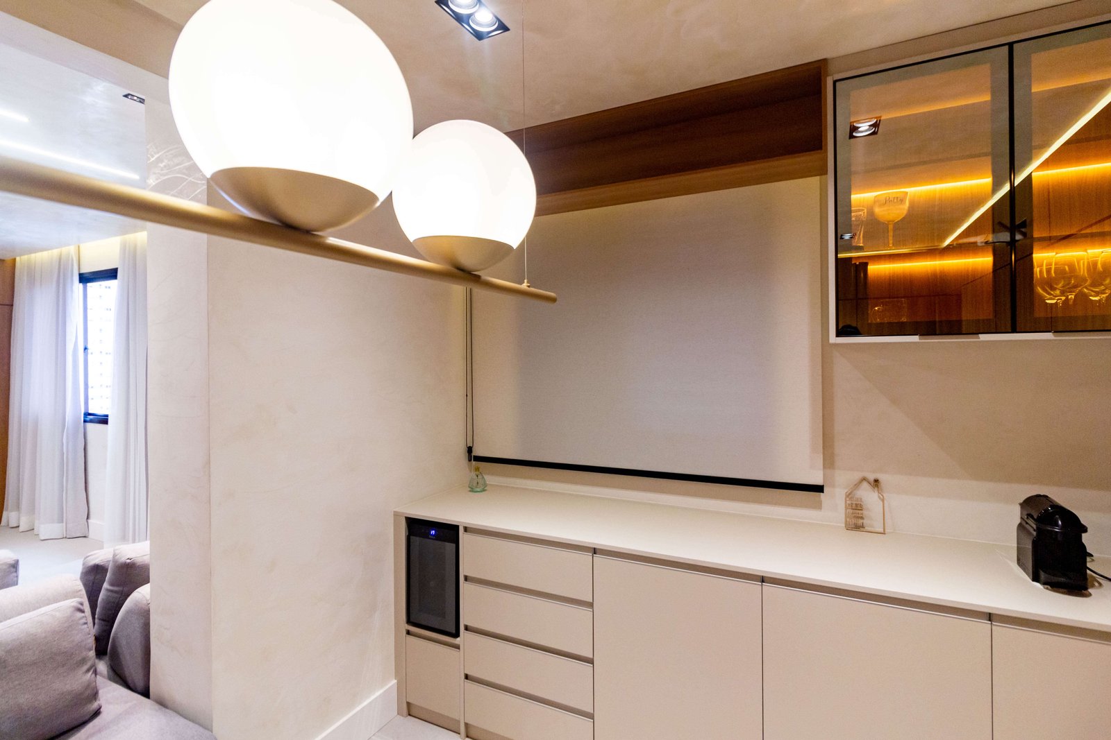 Um cantinho do café ou bar com armários inferiores na cor bege, adega climatizada preta embutida e um pendente moderno dourado com dois globos brancos.