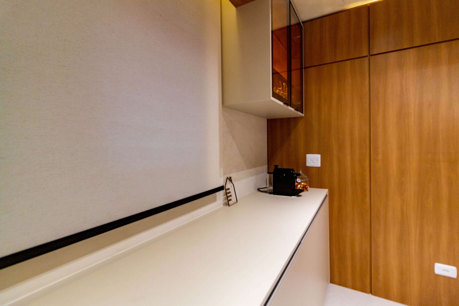 Outro ângulo da bancada, agora exibindo uma cafeteira de cápsulas sobre o móvel e uma parede lateral revestida inteiramente em painel de madeira.