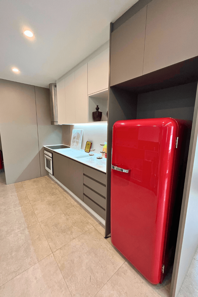 Cozinha linear moderna com armários inferiores cinza-bege e superiores brancos. O destaque é uma geladeira retrô vermelha vibrante ao lado da torre de marcenaria.
