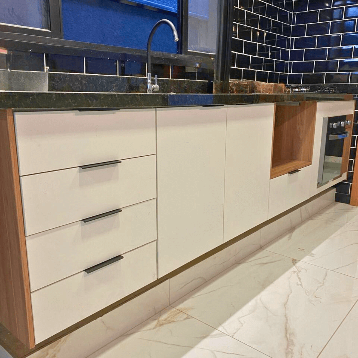 Detalhe de um balcão com gavetões brancos e puxadores pretos discretos. Há um nicho lateral em madeira e, ao fundo, uma parede revestida com azulejos estilo 'metrô' (subway tile) na cor azul marinho escuro.