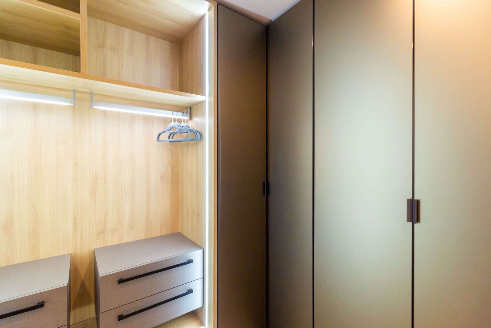 Closet planejado de canto combinando nichos abertos em madeira iluminada com LED e armários fechados com portas escuras foscas.