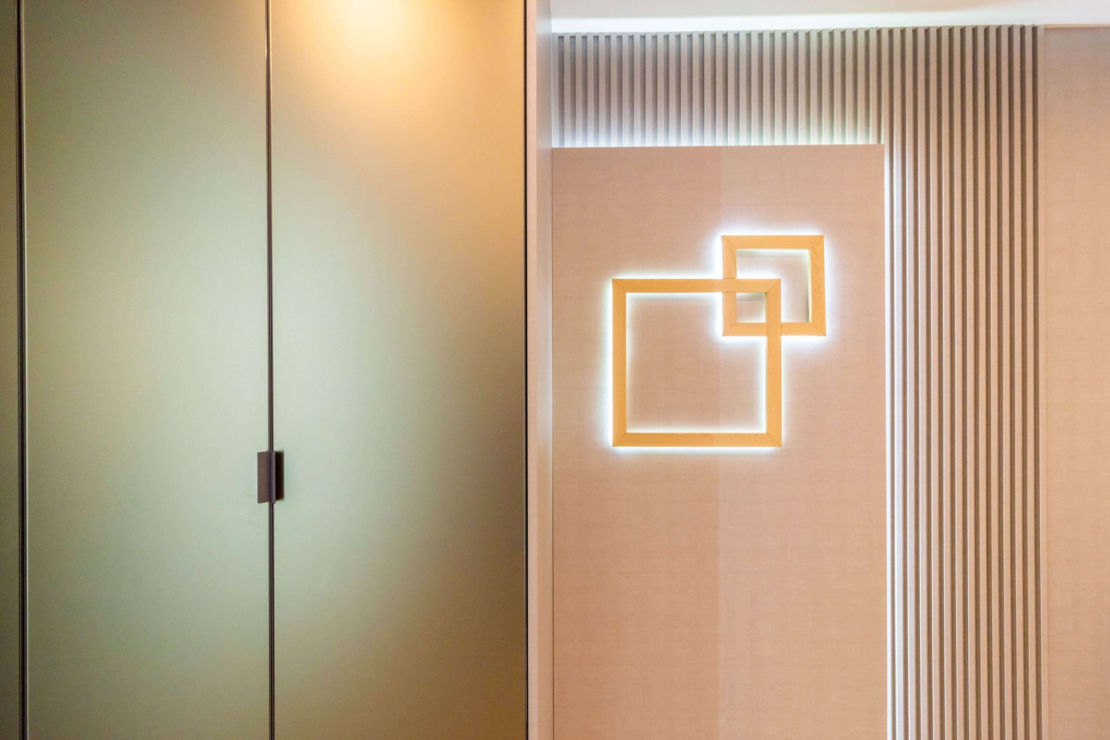 Close-up artístico de um detalhe de marcenaria de alto padrão. À direita, um painel de parede ripado na cor bege serve de fundo para uma luminária de parede (arandela) moderna feita de perfis de LED, formando dois quadrados de luz entrelaçados. À esquerda, vê-se parte de um armário com porta de acabamento liso e acetinado (vidro ou laca) e puxador vertical escuro.