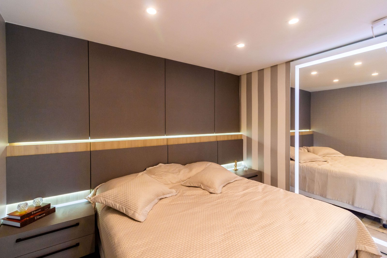 Vista lateral da cama, destacando a iluminação suave do nicho da cabeceira e o reflexo do ambiente na porta de espelho do guarda-roupa ao fundo.