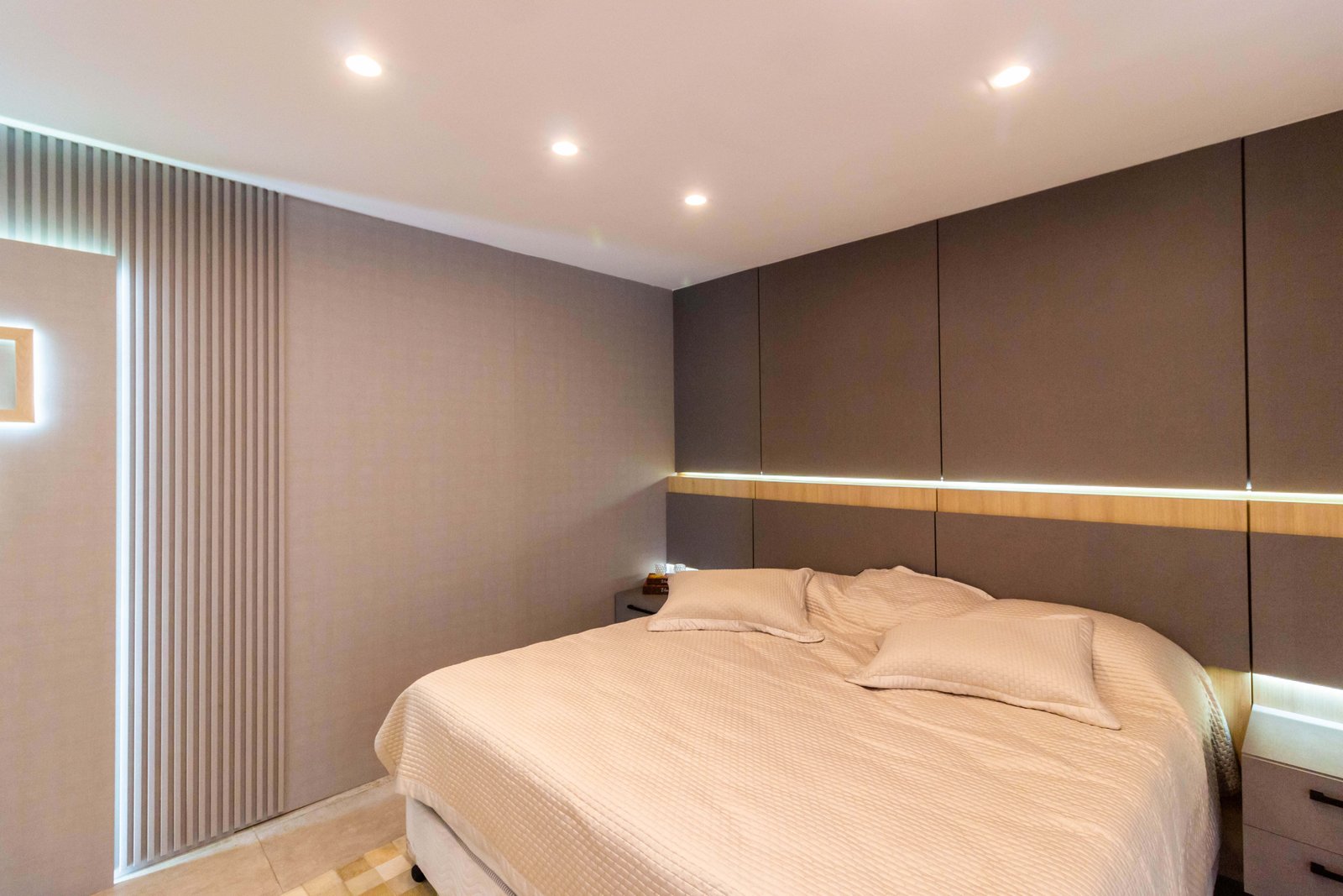 Quarto de casal sofisticado com cabeceira de painéis estofados cinza escuro indo até o teto, cortada por um nicho iluminado. À esquerda, detalhe de um painel ripado.