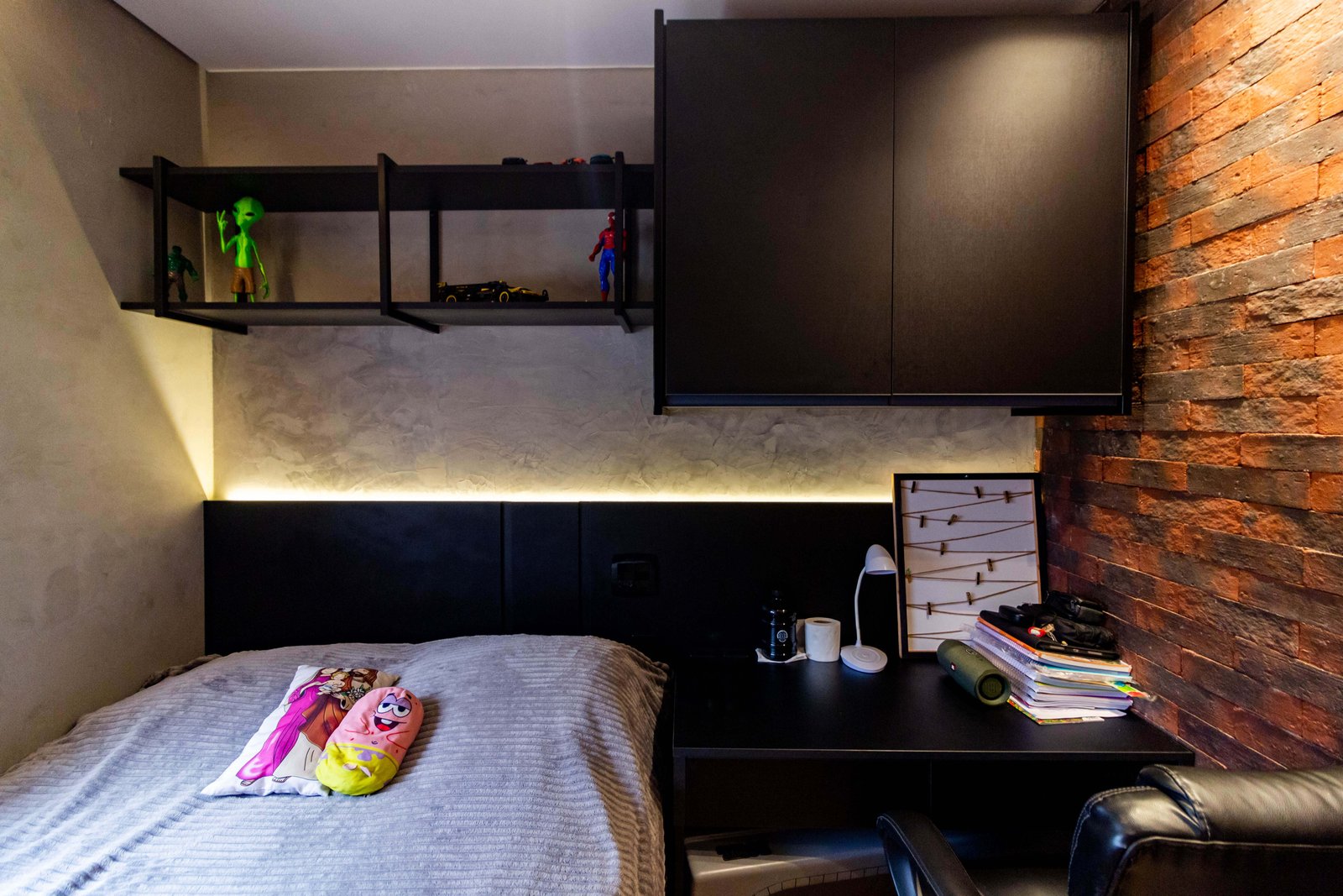 Um quarto juvenil ou 'gamer' com móveis pretos. Vê-se uma escrivaninha, prateleiras aéreas com bonecos de ação e uma cabeceira escura com fita de LED, criando um ambiente moderno e despojado.