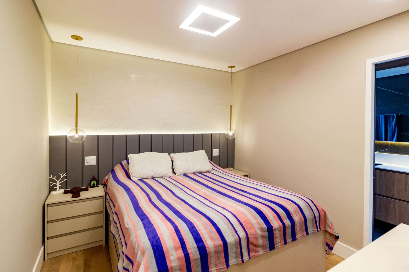 Quarto de hóspedes ou solteiro com uma cama de colcha listrada colorida. A cabeceira é cinza com gomos verticais e há pendentes delicados dourados descendo sobre as mesas de cabeceira.