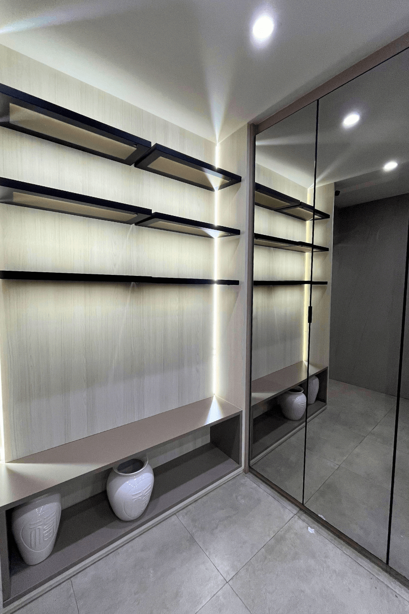 Uma área de vestiário ou closet aberta, com prateleiras de madeira clara com bordas pretas e iluminação de LED vertical. O ambiente conta com grandes espelhos que ampliam o espaço.