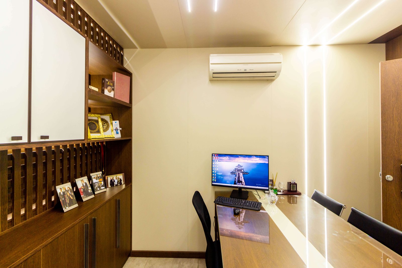 Estação de trabalho executiva com estante de madeira iluminada, mesa de vidro com computador e parede com detalhe em LED vertical.