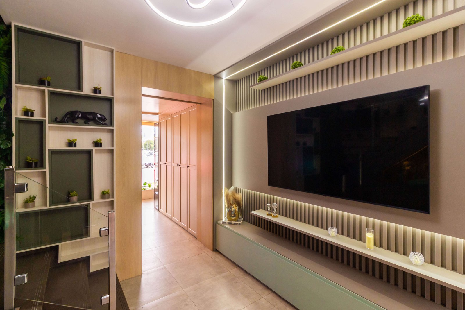 O mesmo ambiente visto de outro ângulo, mostrando a integração da sala de TV com um corredor que possui armários planejados (roupeiro) em madeira clara do chão ao teto.