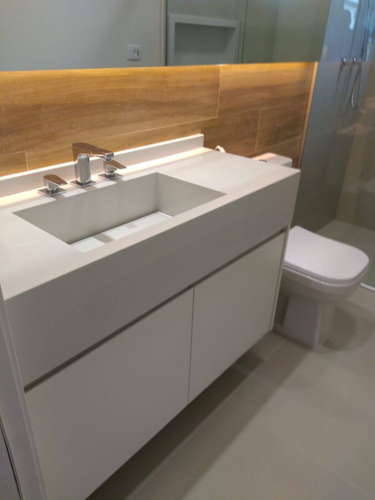 Um banheiro moderno mostrando uma bancada de pedra branca com cuba esculpida, torneira cromada e gabinete branco. Ao fundo, um revestimento na parede que imita madeira com iluminação de LED no espelho.