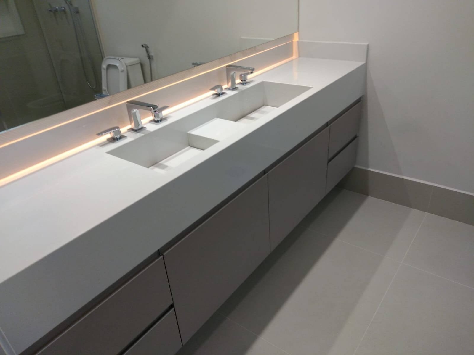 Um banheiro moderno com uma bancada longa em pedra branca e cuba dupla esculpida. Abaixo, armários em tom de cinza quente (greige) e, acima, um espelho amplo com iluminação de LED embutida nas bordas.