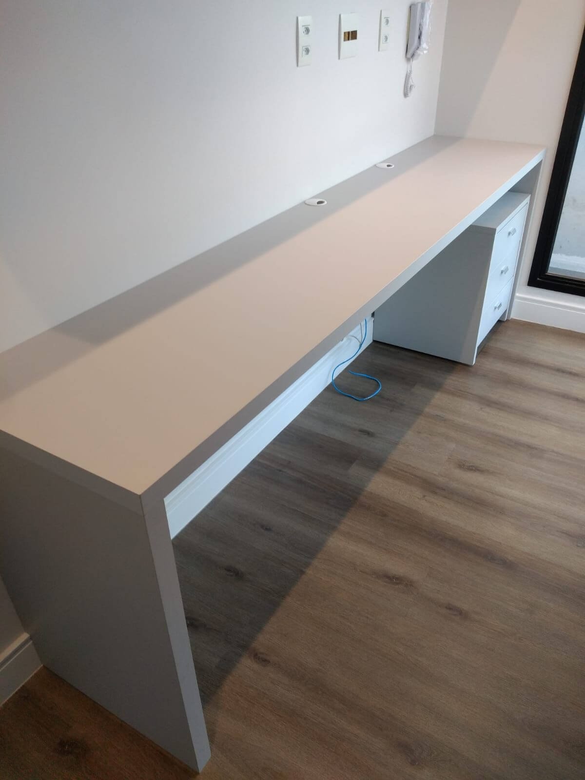 Uma bancada de estudos ou home office longa e minimalista em tom bege claro, fixada na parede, com furos para passagem de cabos. O piso é de madeira escura.