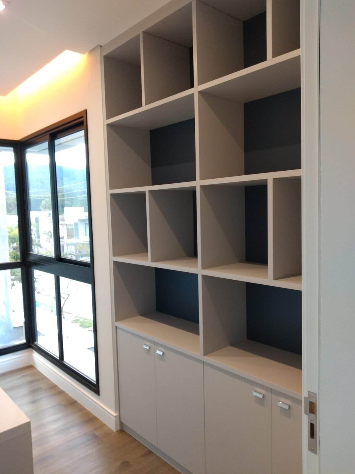 Uma estante alta planejada ao lado de uma grande janela de vidro com esquadria preta. O móvel possui nichos abertos na parte superior (com o fundo em tom azul-marinho ou cinza escuro) e armários fechados na base.